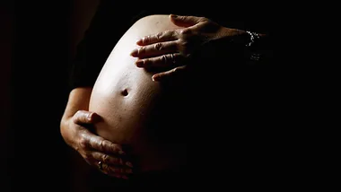 Pregnant woman cradling her bare belly with hands against a dark background.