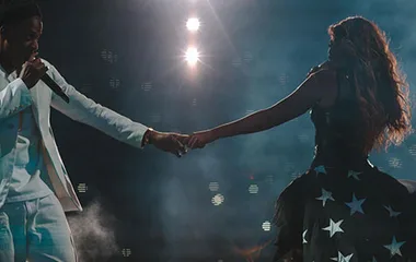 A couple holding hands on stage, illuminated by spotlights, with one dressed in white and the other in a star-patterned outfit.