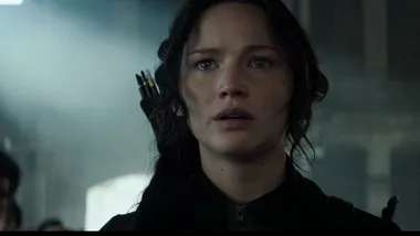 Katniss Everdeen looks determined with arrows on her back in a dimly lit room, from "The Hunger Games: Mockingjay Part 1".