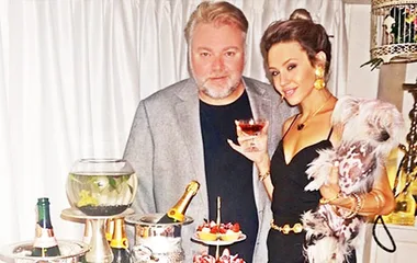 Man and woman posing at a party with drinks and a small dog, standing beside a table with champagne and dessert.