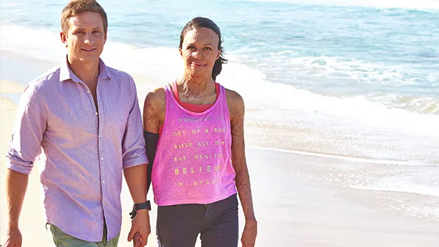 Man and woman walking together on a sunny beach; woman wearing a bright tank top.