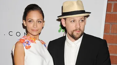 A man in a suit and hat stands beside a woman in a floral dress at an event, smiling at the camera.