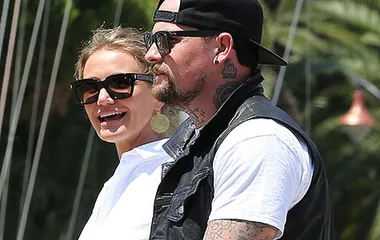 Two people, wearing sunglasses, smiling outdoors. One person has tattoos and a backward cap.