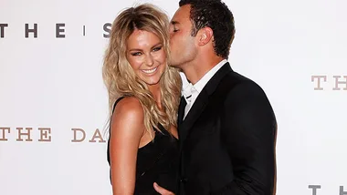 Couple at an event, woman in black dress smiling as man in suit kisses her cheek.