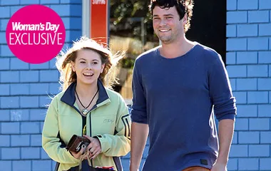 A young woman and man smiling, walking outdoors, she holds a phone; caption reads “Woman’s Day Exclusive.”