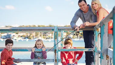 A family of five enjoys fishing on a dock, smiling with a scenic waterfront backdrop.