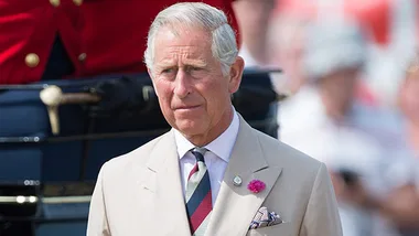 A well-dressed man stands outdoors at a formal event, with others in the background.