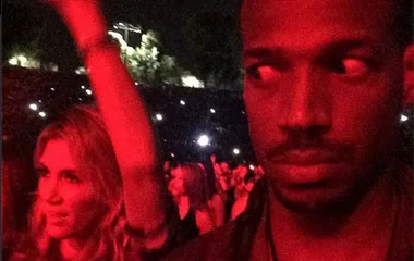 Man at concert looking to his side, woman cheering in background, red lighting, outdoor concert setting.