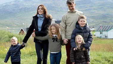 Family of six, warmly dressed, standing outside in a scenic mountainous area with grass and houses in the background.