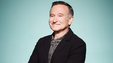 A smiling man in a dark jacket with a light teal background.