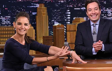 Katie Holmes smiling and gesturing with a man on a talk show set, cityscape backdrop behind them.