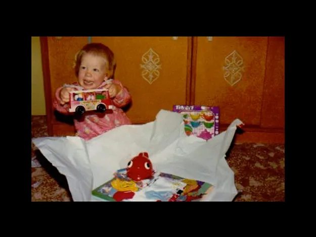 A happy baby holding a toy car, surrounded by wrapping paper and another toy on a carpeted floor.