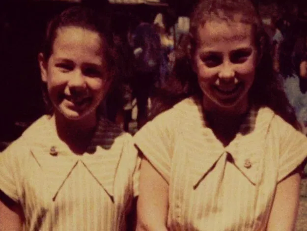 Two girls smiling in vintage-style dresses, standing close together outdoors.