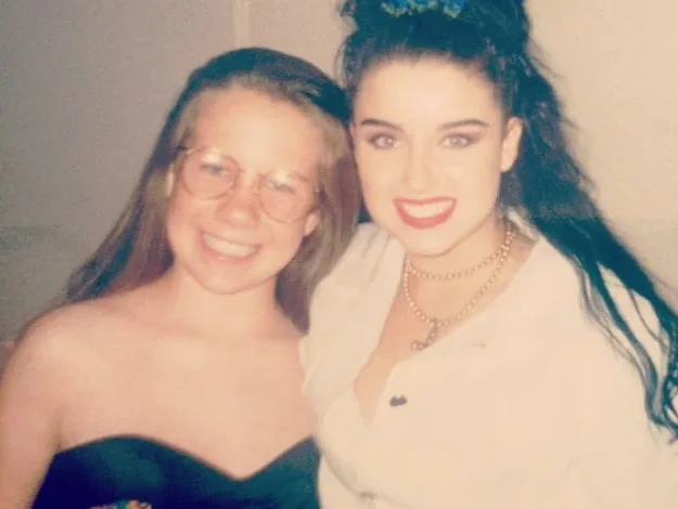 Two women smiling, one wearing glasses and a black dress, the other with dark hair in a white shirt and gold necklace.