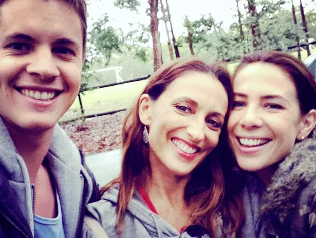 Three people smiling outdoors, taking a selfie in a wooded area with trees in the background.