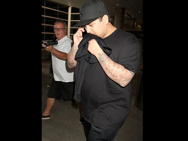 Person in a black shirt with tattoos covers face while being photographed at an airport.