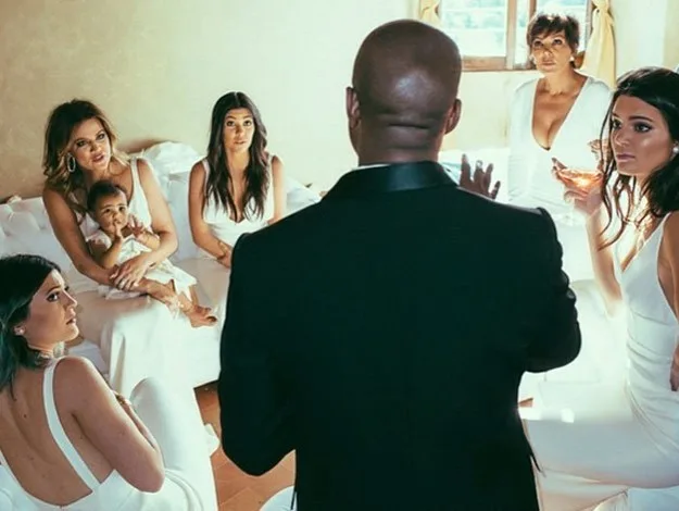A man in a suit talks to women dressed in white seated on a sofa, one holding a baby, in a softly lit room.