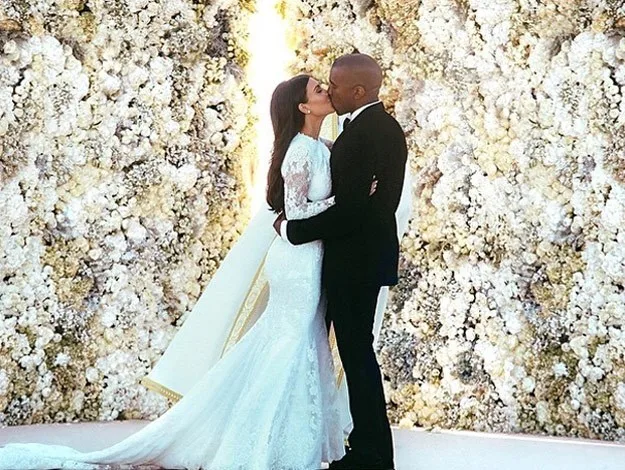 Bride in lace gown and groom in tuxedo kiss before a wall of white flowers.