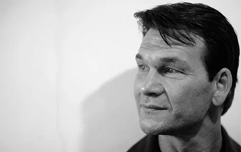 A black and white portrait of Patrick Swayze gazing thoughtfully to the side, with a neutral expression on his face.