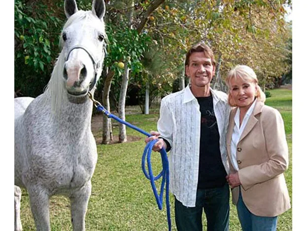 Man and woman standing with a white horse in a garden, smiling at the camera.