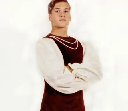 Young man in medieval-style costume with a red tunic and white sleeves, arms crossed, gazing confidently.