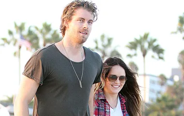 Two people walking outdoors in a sunny setting, with palm trees and a US flag in the background.