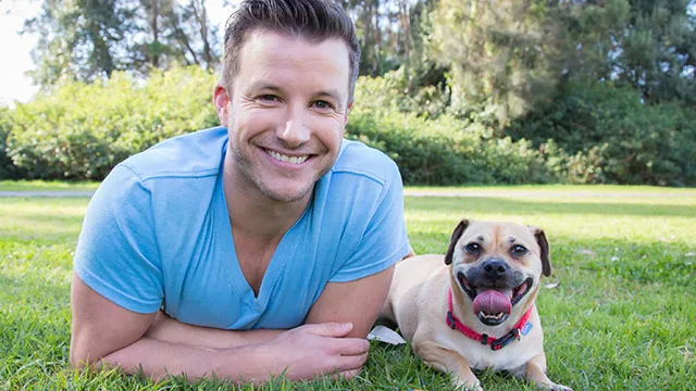 A person in a blue shirt lying on grass next to a happy dog with trees in the background.