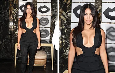 Kim Kardashian wearing a revealing black jumpsuit, standing indoors in front of artwork with lip designs.