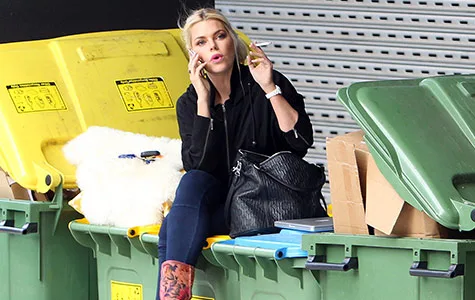 Sophie Monk is sitting on a garbage bin, talking on the phone, holding a cigarette, with bags and boxes around.