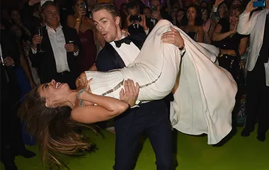 Dancing pair at a formal event; woman in white dress, man in black tuxedo, surrounded by a lively crowd.