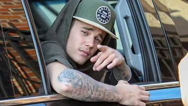 A person in a green cap and hoodie leans out of a vehicle window, pointing at the camera.