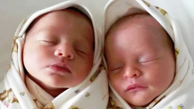 Twin newborns wrapped in patterned blankets, peacefully sleeping side by side.