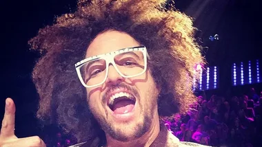 A person with curly hair and white glasses smiles widely, pointing upwards in a vibrant, lit-up setting.