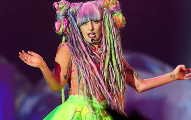Lady Gaga performs on stage in Australia wearing a colorful, eccentric outfit and rainbow hair, singing passionately.