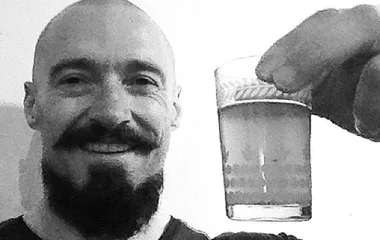 Man with a shaved head and beard holding a small glass, smiling at the camera.