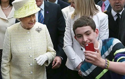 A young person takes a selfie with an elderly woman in a yellow outfit and hat, surrounded by a crowd.