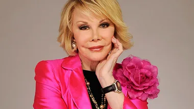 A woman in a bright pink jacket with a flower detail poses thoughtfully, hand on cheek, against a neutral background.