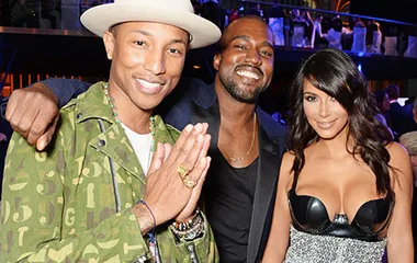 Pharrell Williams, Kanye West, and Kim Kardashian smiling together at an event, with Pharrell in a green jacket and hat.