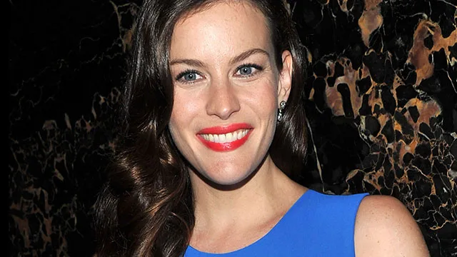 Liv Tyler smiling in a blue dress with curled hair against a dark, textured background.
