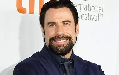 John Travolta in a suit with a thick beard, smiling at a film festival event.
