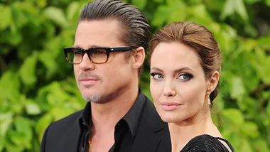A man and woman in formal attire stand together with greenery in the background.
