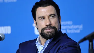 John Travolta at a press conference, wearing a blue suit and white shirt, against a blue background.