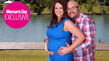 Couple smiling by a lake, woman in blue dress showing baby bump, "Woman's Day Exclusive" logo visible.