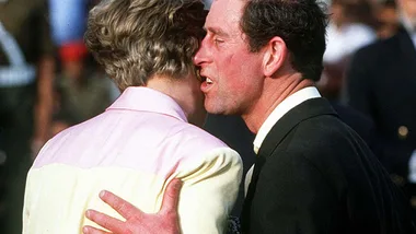 Prince in suit attempts to kiss a woman in a pink and cream outfit, facing away, amidst a crowd.