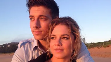 Couple embracing on a beach at sunset, gazing into the distance with serene expressions.