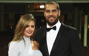 A couple poses elegantly on the red carpet; the man in a tuxedo and the woman in a light gray dress, both smiling.