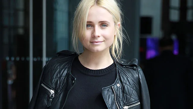 Blonde woman in black leather jacket smiling softly, standing indoors.