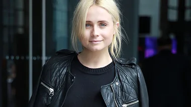 Blonde woman in black leather jacket smiling softly, standing indoors.