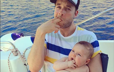 "Man in striped shirt and cap with baby, both mimicking finger-in-mouth gesture, sitting on a boat with sea in background."