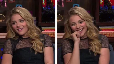 Chloe Grace Moretz smiling and laughing during a talk show interview, wearing a black dress with wavy blonde hair.